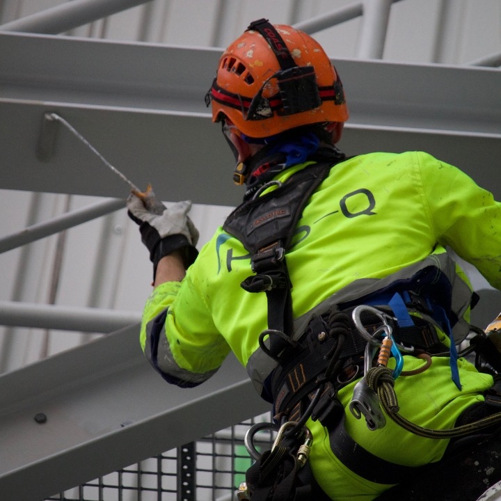 Pracownik w kasku i odzieży ochronnej, zabezpieczony linami, maluje metalową konstrukcję na wysokości. Widok z dołu na plecy, z elementami rusztowania w kadrze.