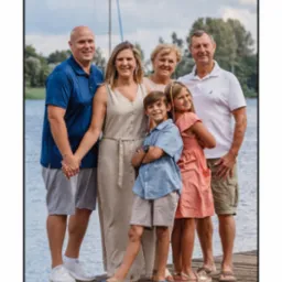 Rodzinna fotografia plenerowa: uśmiechnięta wielopokoleniowa rodzina pozuje na drewnianym pomoście nad jeziorem, w tle widać maszt żaglówki i zieleń drzew.