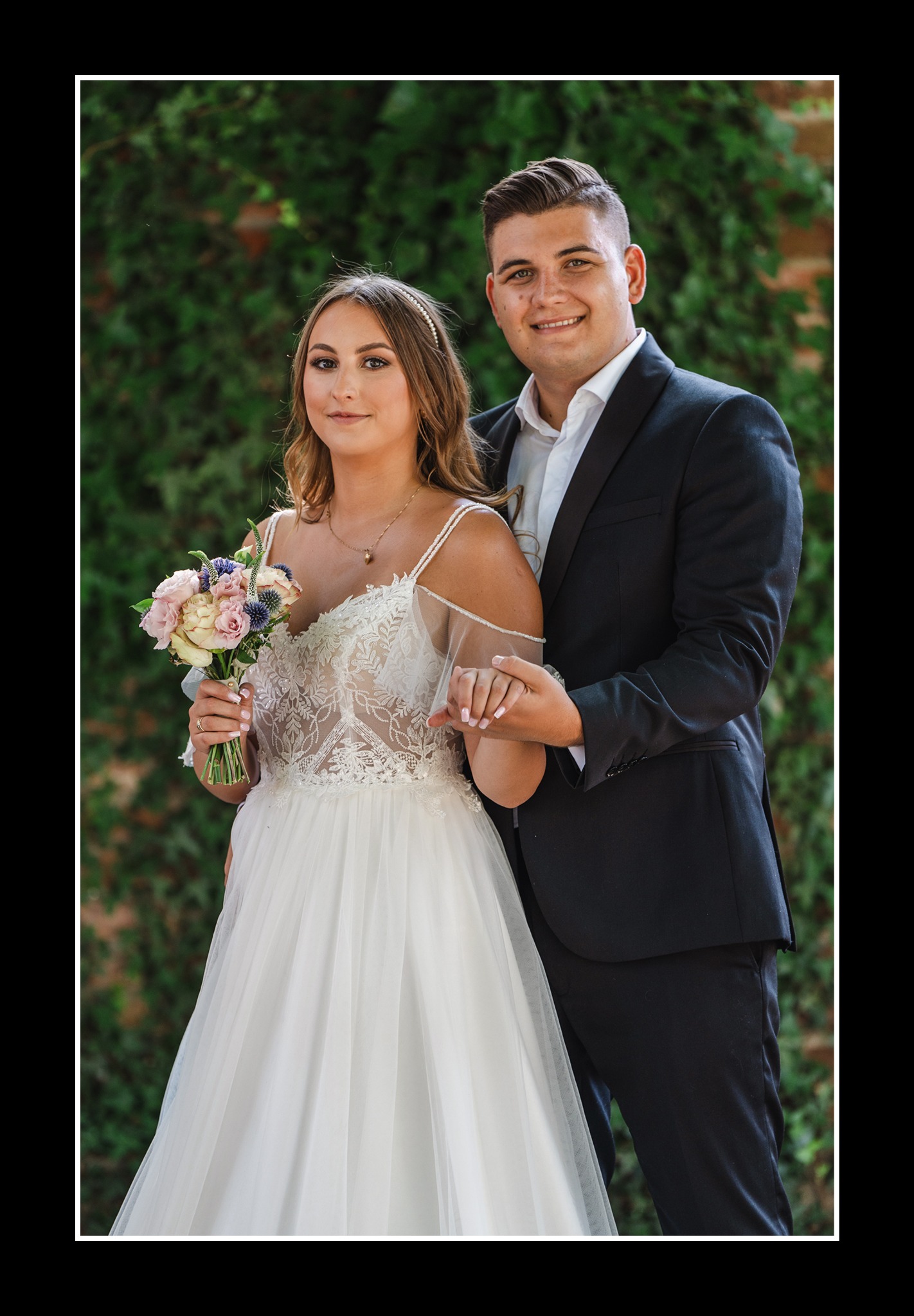 Uśmiechnięta para młoda w plenerze: panna młoda w koronkowej sukni trzyma bukiet, pan młody w czarnym garniturze. Tło stanowi ściana porośnięta bluszczem.