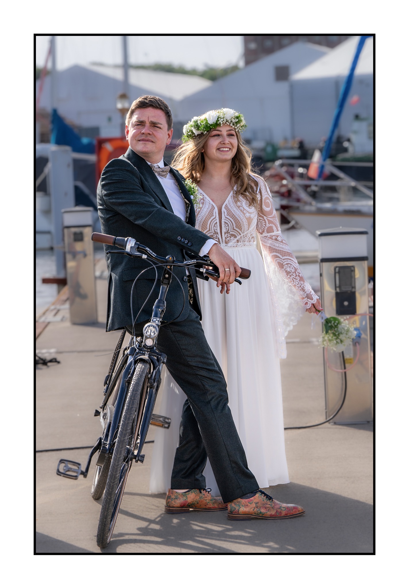 Para młoda na tle portu: pan w ciemnozielonym garniturze oparty o rower, panna młoda w białej sukni z wiankiem na głowie, trzyma bukiet.