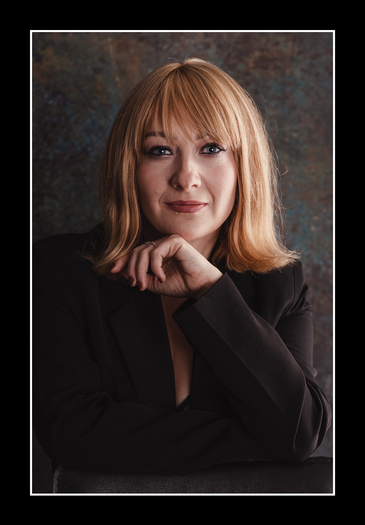 Portret kobiety z blond włosami, w ciemnej marynarce, opierającej brodę na dłoni, na tle abstrakcyjnej tekstury. Subtelny makijaż i wyraz oczu budują profesjonalny wizerunek.