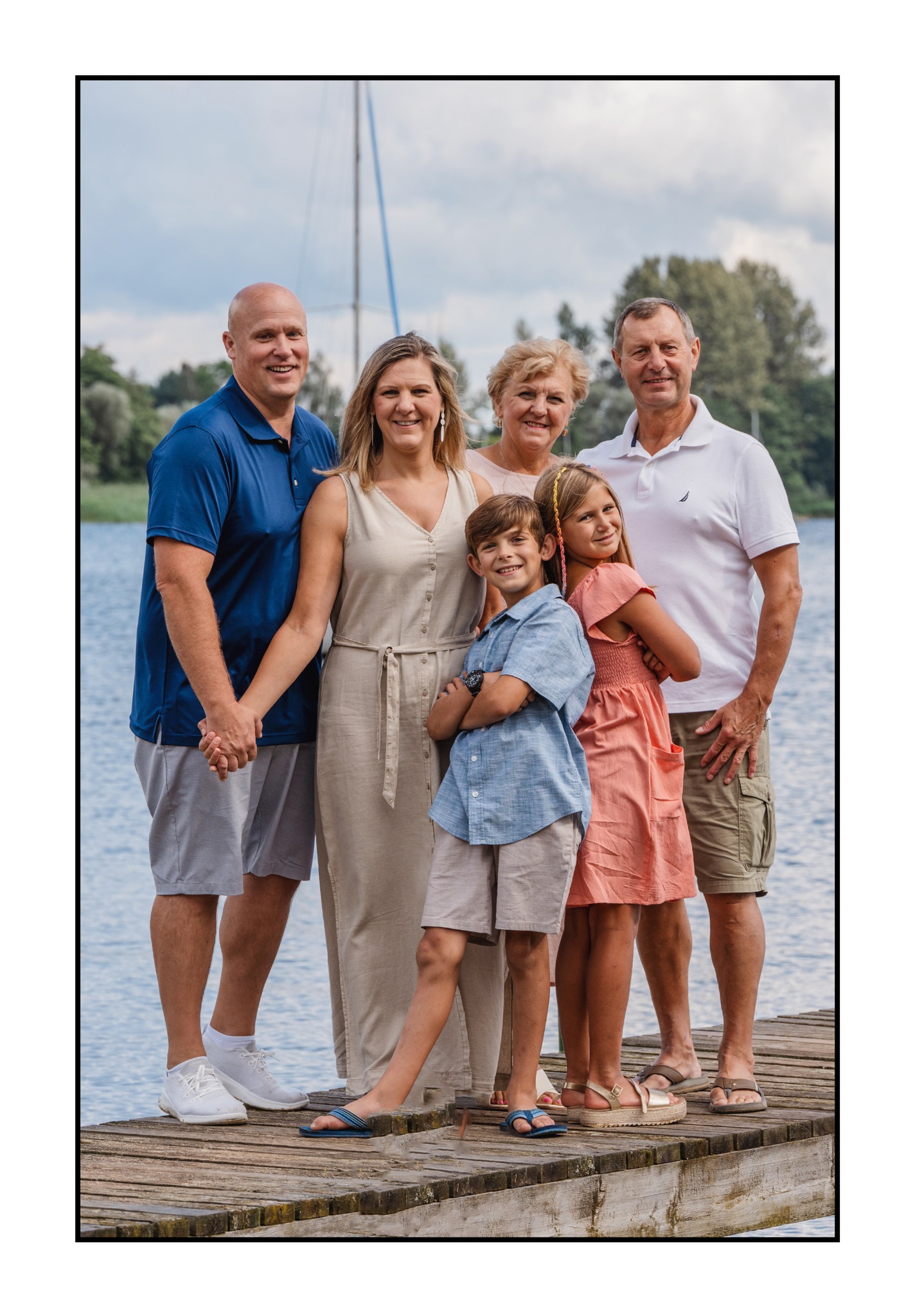 Rodzinna fotografia plenerowa: uśmiechnięta wielopokoleniowa rodzina pozuje na drewnianym pomoście nad jeziorem, w tle widać maszt żaglówki i zieleń drzew.