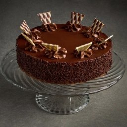 ZŁOTY KLUCZ SPÓŁKA Z OGRANICZONĄ ODPOWIEDZIALNOŚCIĄ - Czekoladowy tort na szklanej paterze, ozdobiony kremem i czekoladowymi dekoracjami, posypany brązową posypką. Gładka polewa, elegancki wygląd.
