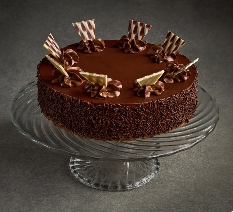 Czekoladowy tort na szklanej paterze, ozdobiony kremem i czekoladowymi dekoracjami, posypany brązową posypką. Gładka polewa, elegancki wygląd.