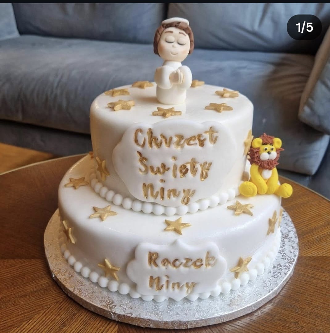 Dwupoziomowy tort z białym lukrem i złotymi gwiazdkami, dedykowany na chrzest i roczek Niny, z figurką aniołka i lwa. Dekoracja na drewnianym stole.