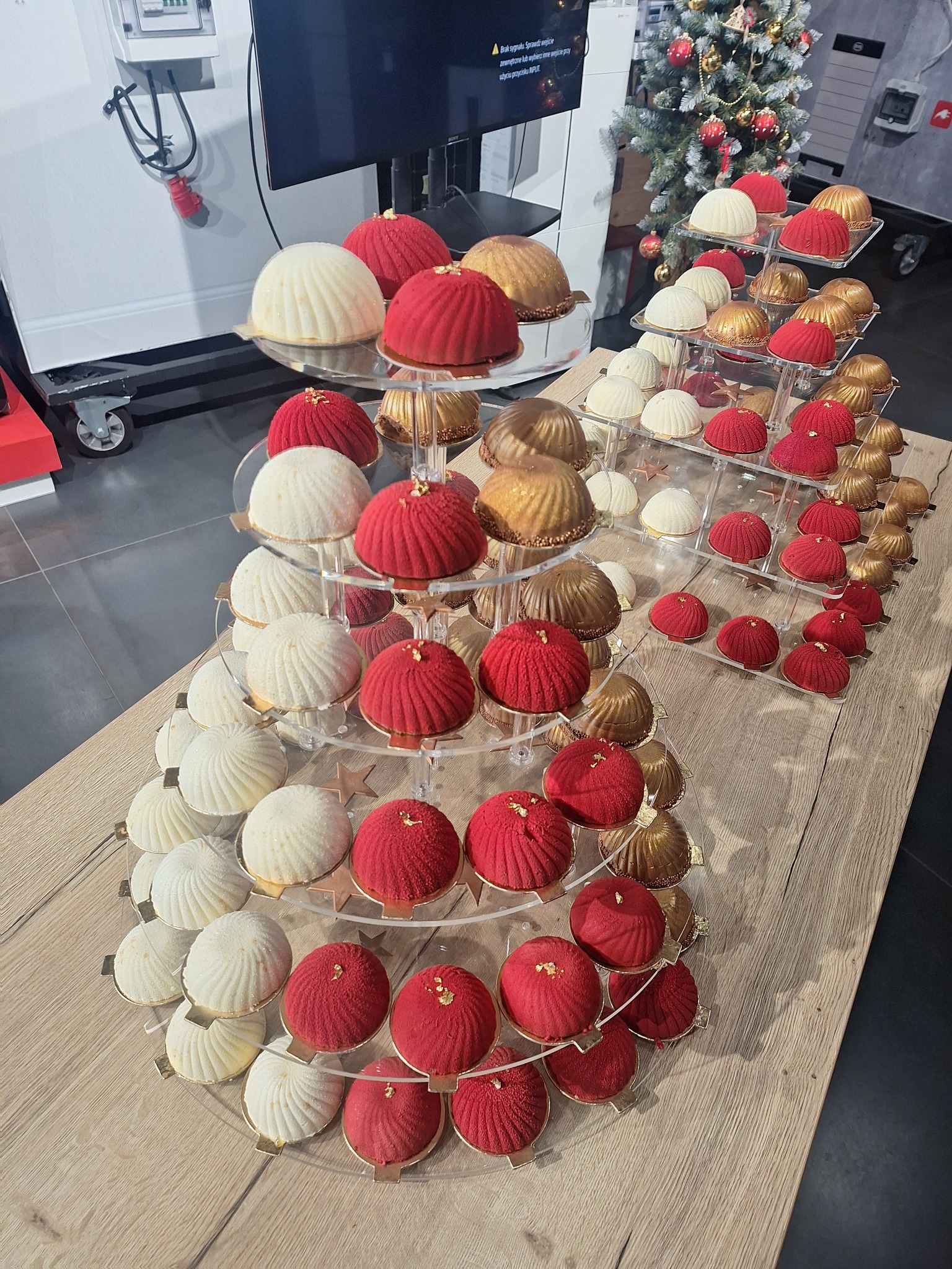 Elegancka ekspozycja deserów w kolorach czerwieni, złota i bieli na wielopoziomowym stojaku z pleksi, w tle choinka świąteczna i sprzęt cateringowy.