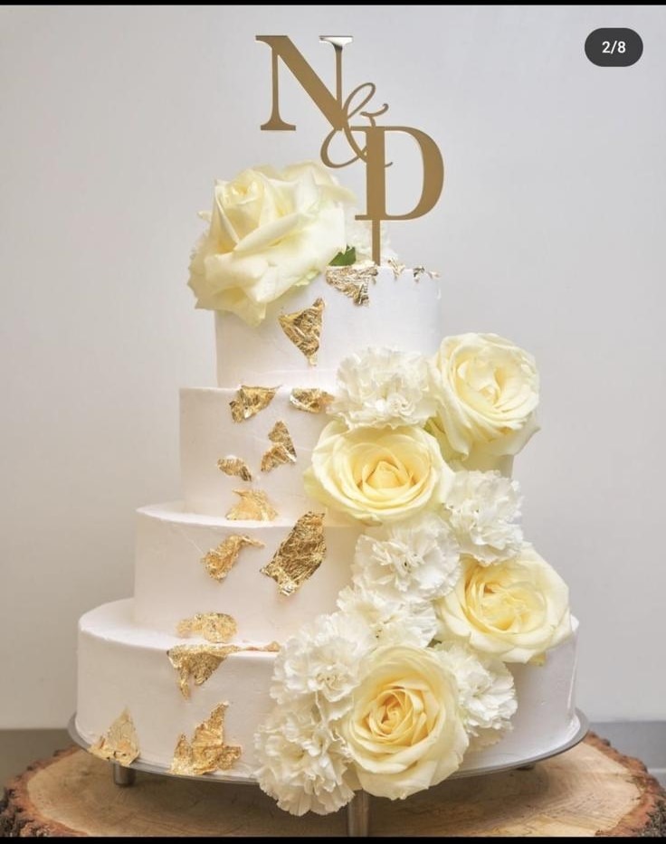 Elegancki tort weselny, czteropiętrowy, z białym kremem, zdobiony złotymi płatkami i kremowymi różami. Na szczycie topper z inicjałami N&D.