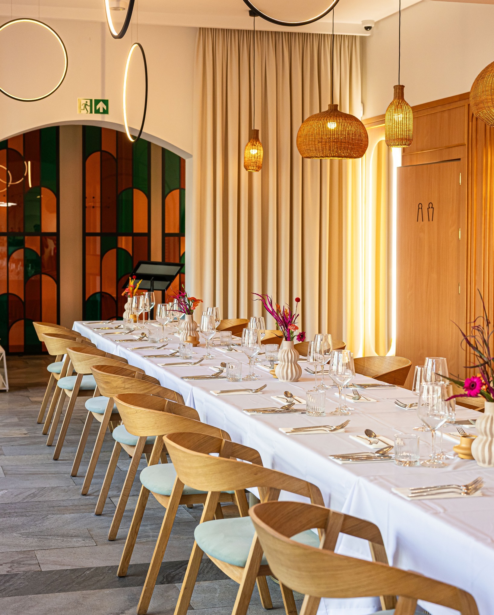 Elegancko nakryty stół z białym obrusem, szklanymi naczyniami i bukietami kwiatów. Krzesła z jasnego drewna. W tle dekoracyjne lampy i wzorzysta ściana.