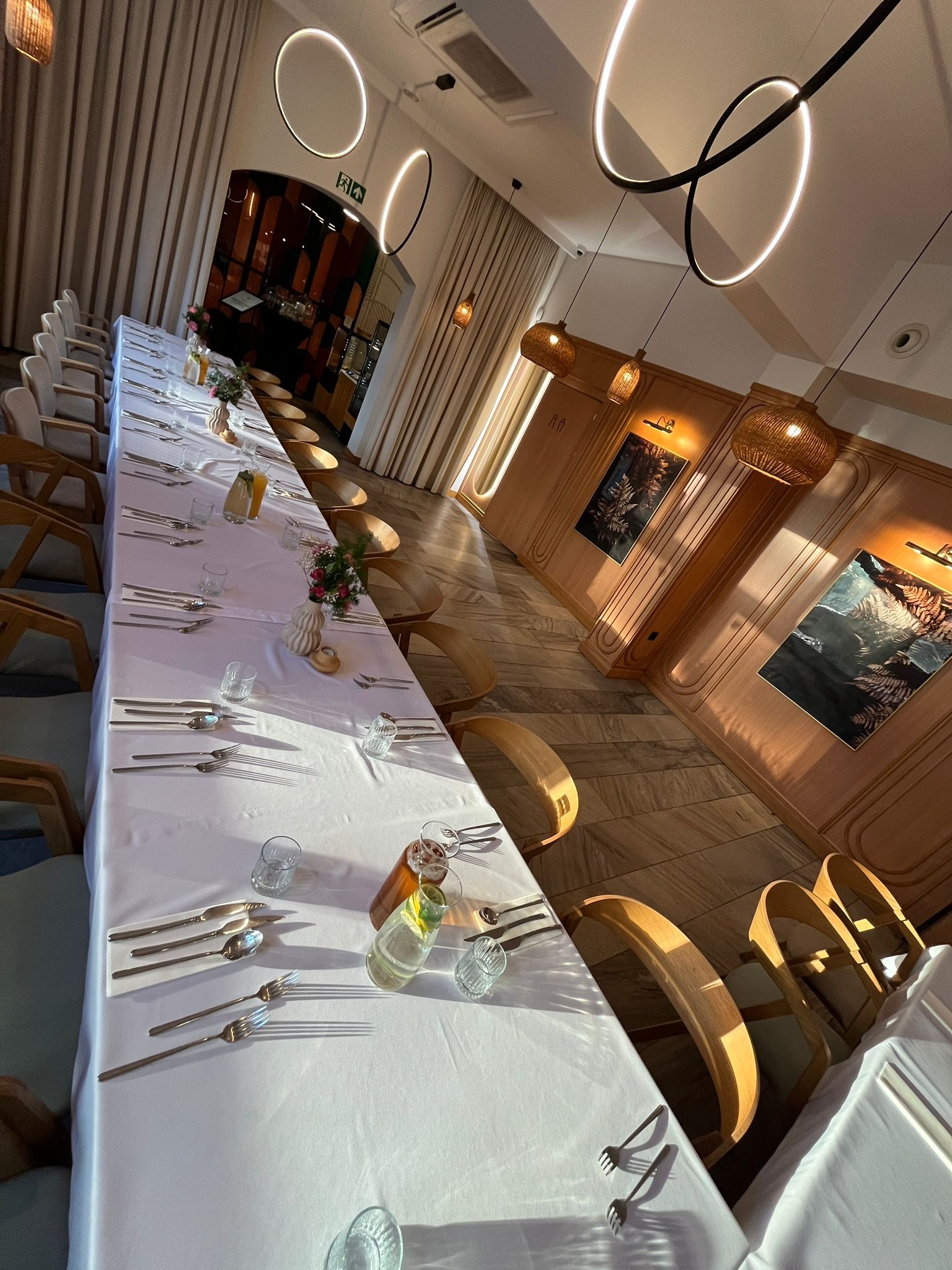 Elegancko nakryty, długi stół z białym obrusem, srebrnymi sztućcami i szklankami, gotowy na przyjęcie w jasnym wnętrzu z drewnianymi akcentami i dekoracyjnym oświetleniem.