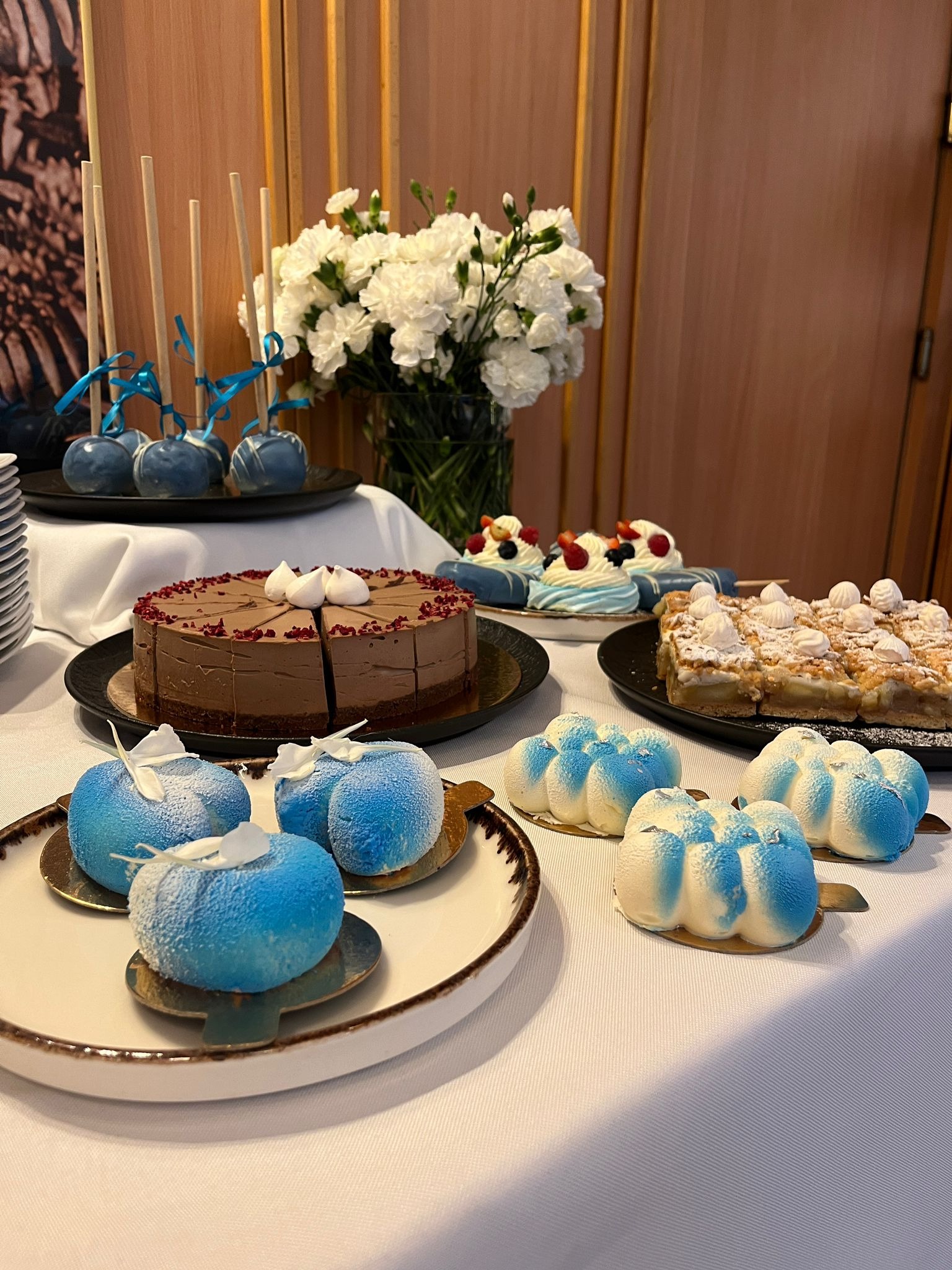 Dekoracyjny stół z różnorodnymi ciastami i deserami w odcieniach błękitu i brązu, udekorowany kwiatami, na białym obrusie i tle drewnianej ściany.