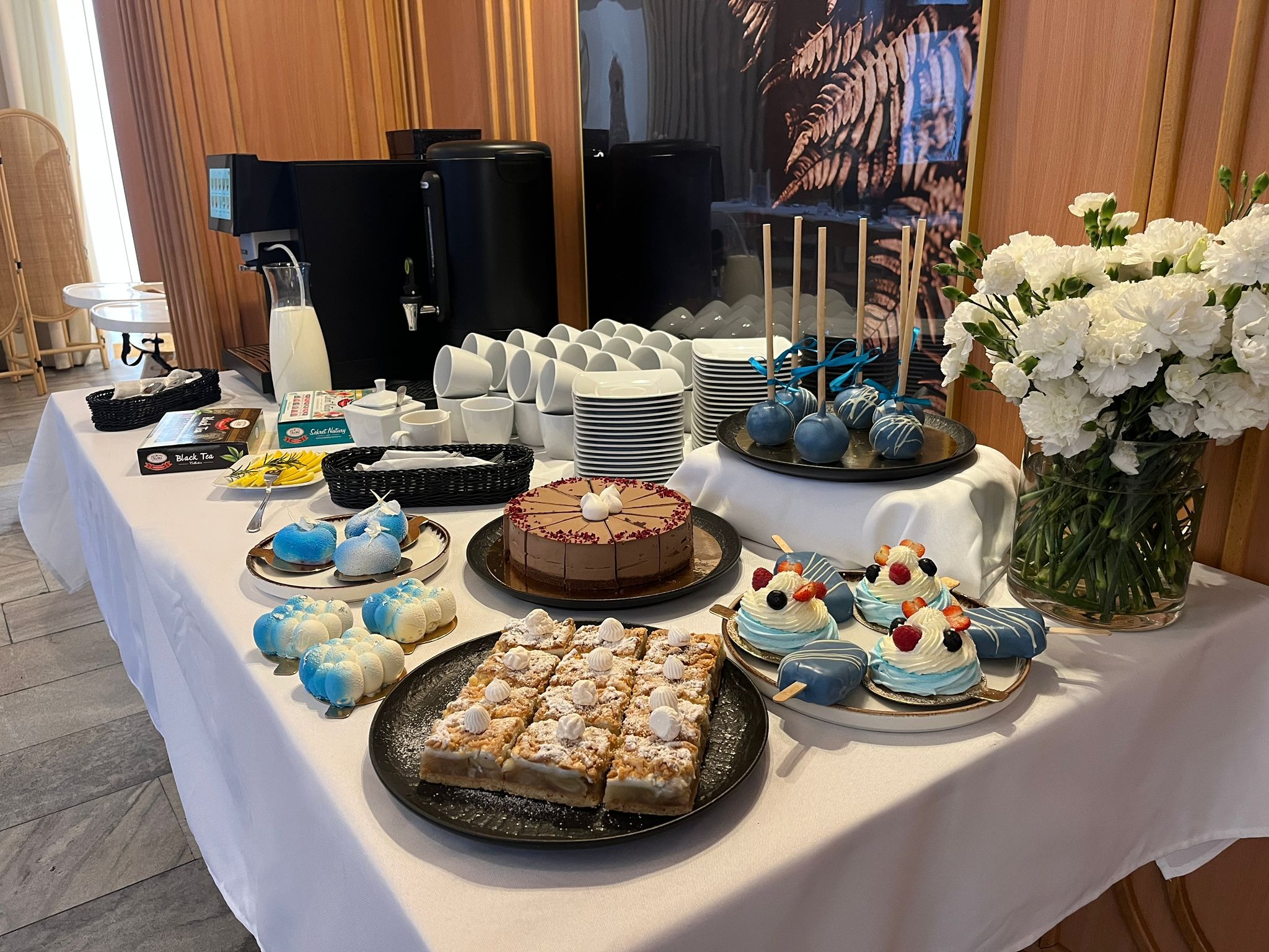 Stół z deserami: tort czekoladowy, ciasto z kruszonką, babeczki z owocami, cake pops, dekoracje kwiatowe i ekspres do kawy. Bufet słodkości.