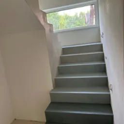 Schody wewnętrzne wykończone szarą, gładką powierzchnią. Widoczne okno z widokiem na zieleń. Minimalistyczny design i jednolite wykończenie ścian.