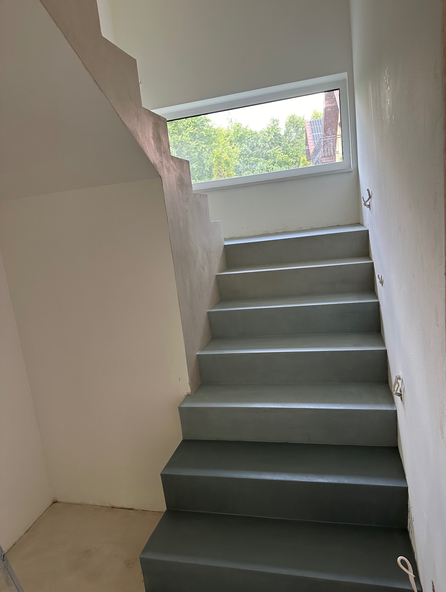 Schody wewnętrzne wykończone szarą, gładką powierzchnią. Widoczne okno z widokiem na zieleń. Minimalistyczny design i jednolite wykończenie ścian.