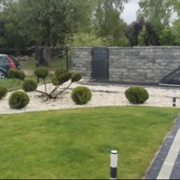Zadbany ogród z równo przystrzyżonym trawnikiem, ozdobnymi krzewami i białym żwirem. W tle ogrodzenie z bramą i szarym murem. Widoczny fragment podjazdu.