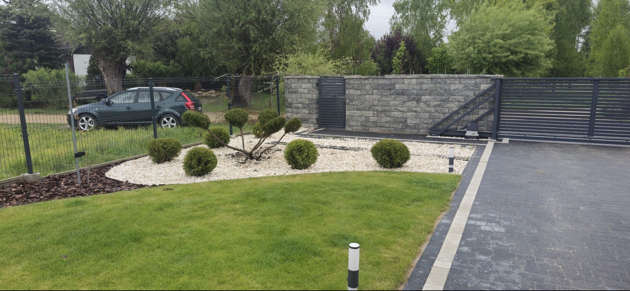 Zadbany ogród z równo przystrzyżonym trawnikiem, ozdobnymi krzewami i białym żwirem. W tle ogrodzenie z bramą i szarym murem. Widoczny fragment podjazdu.