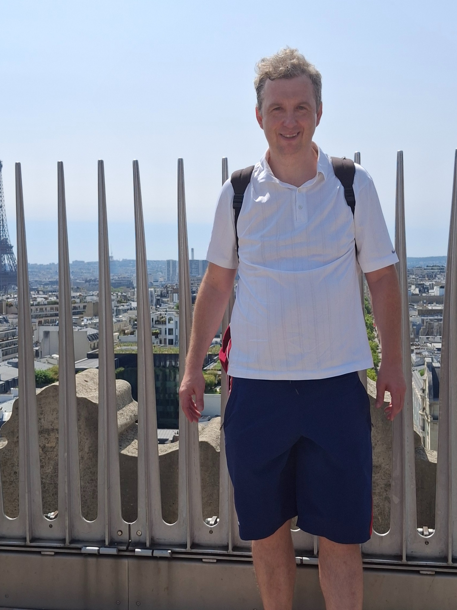 Uśmiechnięty mężczyzna w białej koszulce polo i granatowych spodenkach, z plecakiem, na tle panoramy Paryża z Wieżą Eiffla w tle.