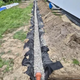 Wykonany drenaż opaskowy przy budynku, widoczna rura drenarska, geowłóknina i wypełnienie żwirowe. Osoba w tle. Ujęcie z góry.