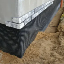 Ochrona fundamentów budynku z użyciem folii kubełkowej, zabezpieczenie przed wilgocią. Widoczne ceglane wykończenie i ziemia wokół fundamentów.