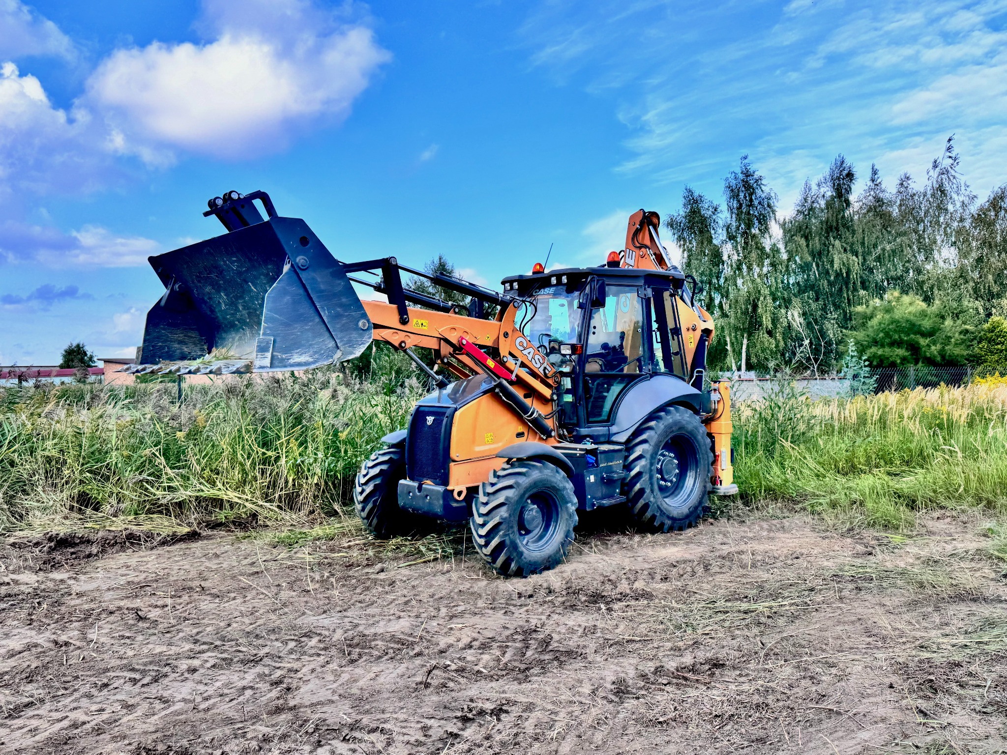Pomarańczowa koparko-ładowarka Case na placu budowy z podniesioną łyżką, na tle niebieskiego nieba i drzew. Widoczne ślady opon na ziemi i trawa.