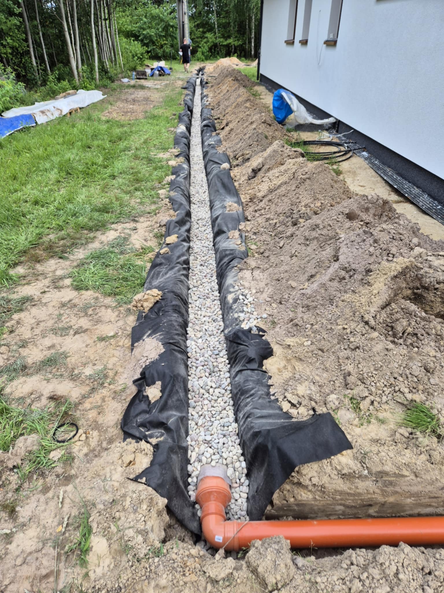 Wykonany drenaż opaskowy przy budynku, widoczna rura drenarska, geowłóknina i wypełnienie żwirowe. Osoba w tle. Ujęcie z góry.