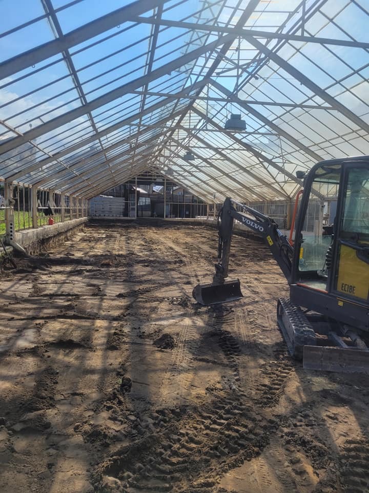 Żółta minikoparka Volvo wewnątrz szklarni z metalową konstrukcją i ziemią przygotowaną pod uprawę. Widoczne ślady opon na ziemi.