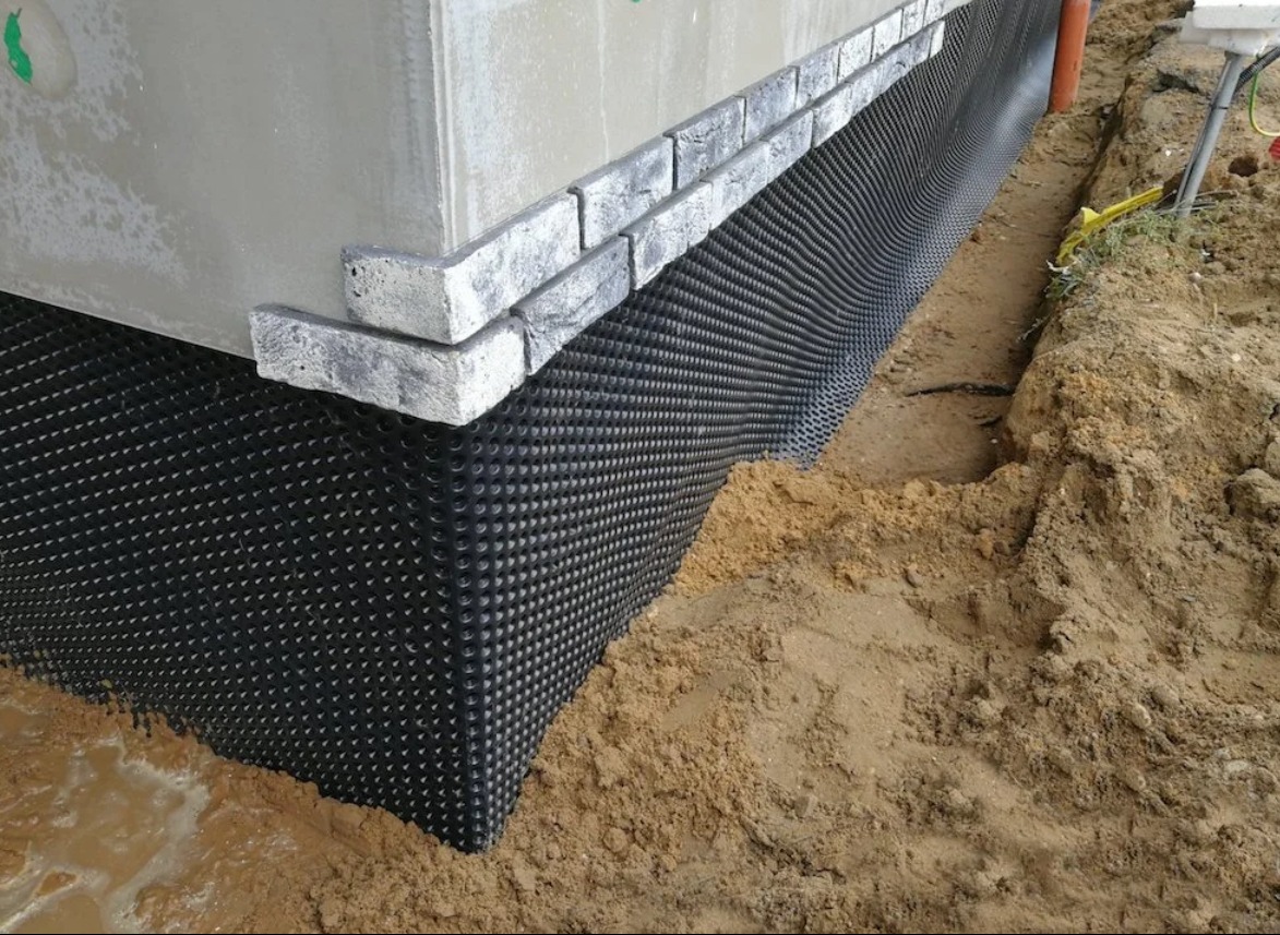 Ochrona fundamentów budynku z użyciem folii kubełkowej, zabezpieczenie przed wilgocią. Widoczne ceglane wykończenie i ziemia wokół fundamentów.