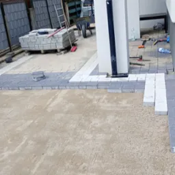 Fragment układania kostki brukowej w odcieniach szarości i bieli na podsypce piaskowej, widoczne narzędzia i materiały budowlane. Praca w toku.