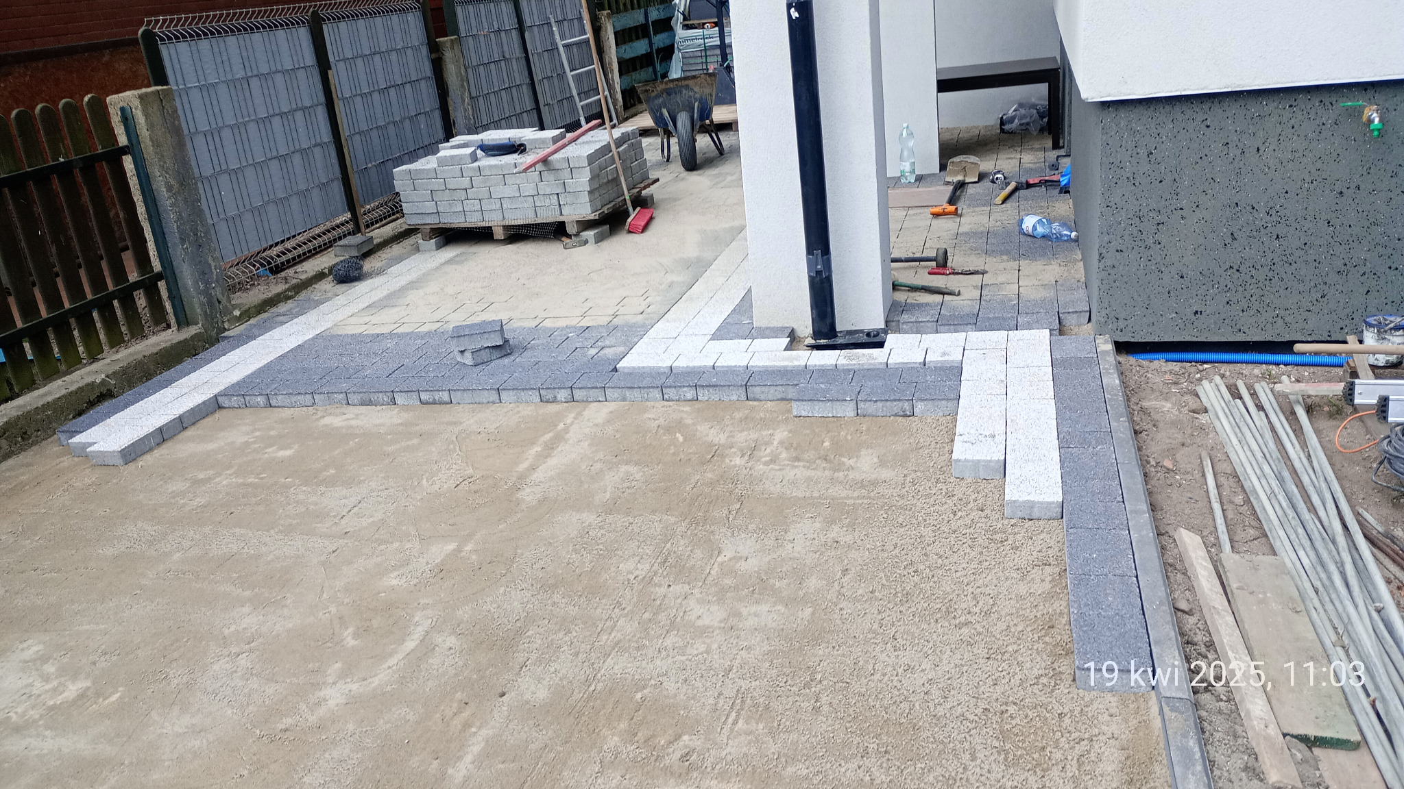 Fragment układania kostki brukowej w odcieniach szarości i bieli na podsypce piaskowej, widoczne narzędzia i materiały budowlane. Praca w toku.