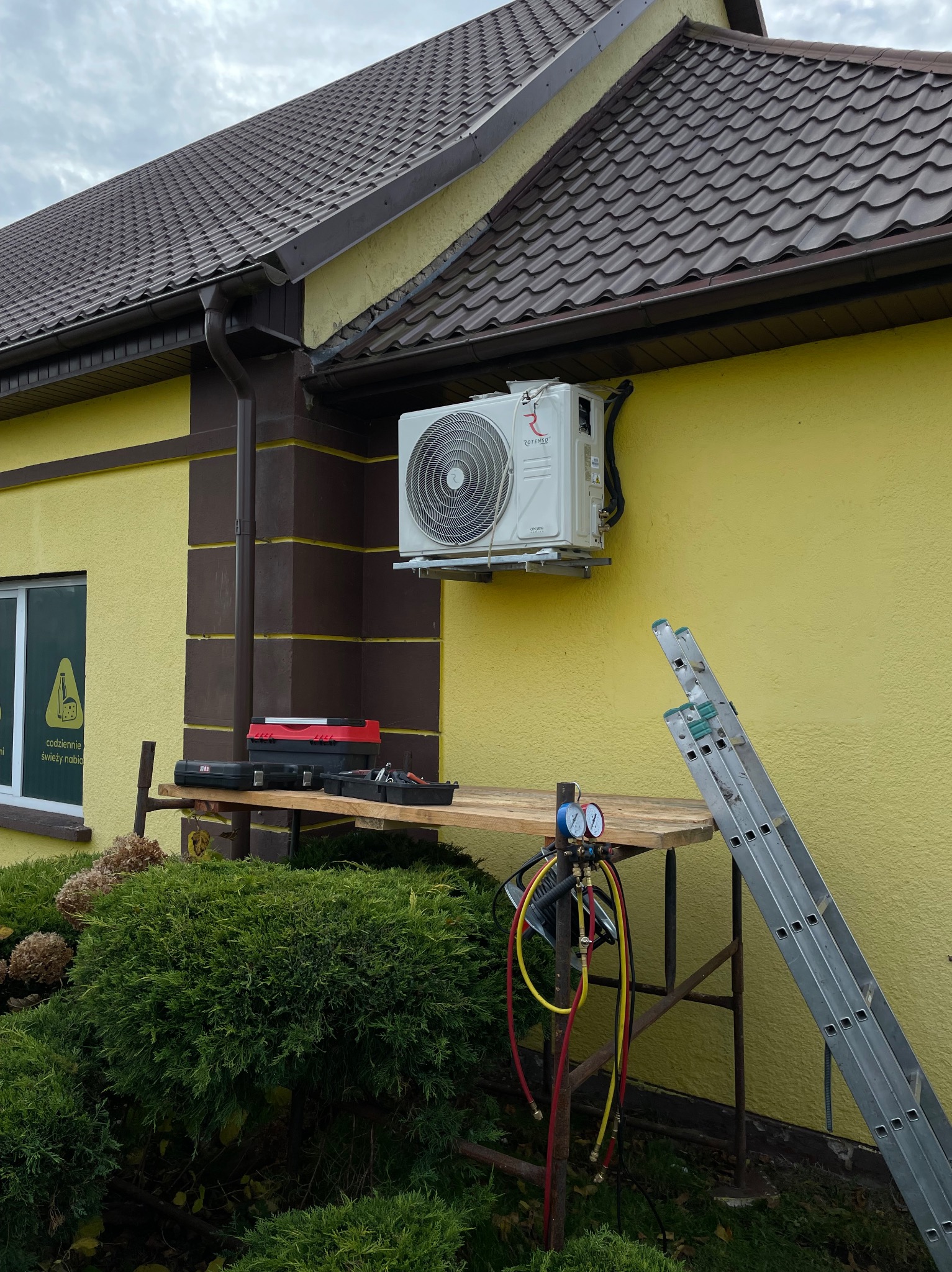 Biała klimatyzacja Rotenso zamontowana na żółtej ścianie domu. Obok drabina i narzędzia na rusztowaniu. Widok na elewację budynku z brązowymi elementami.