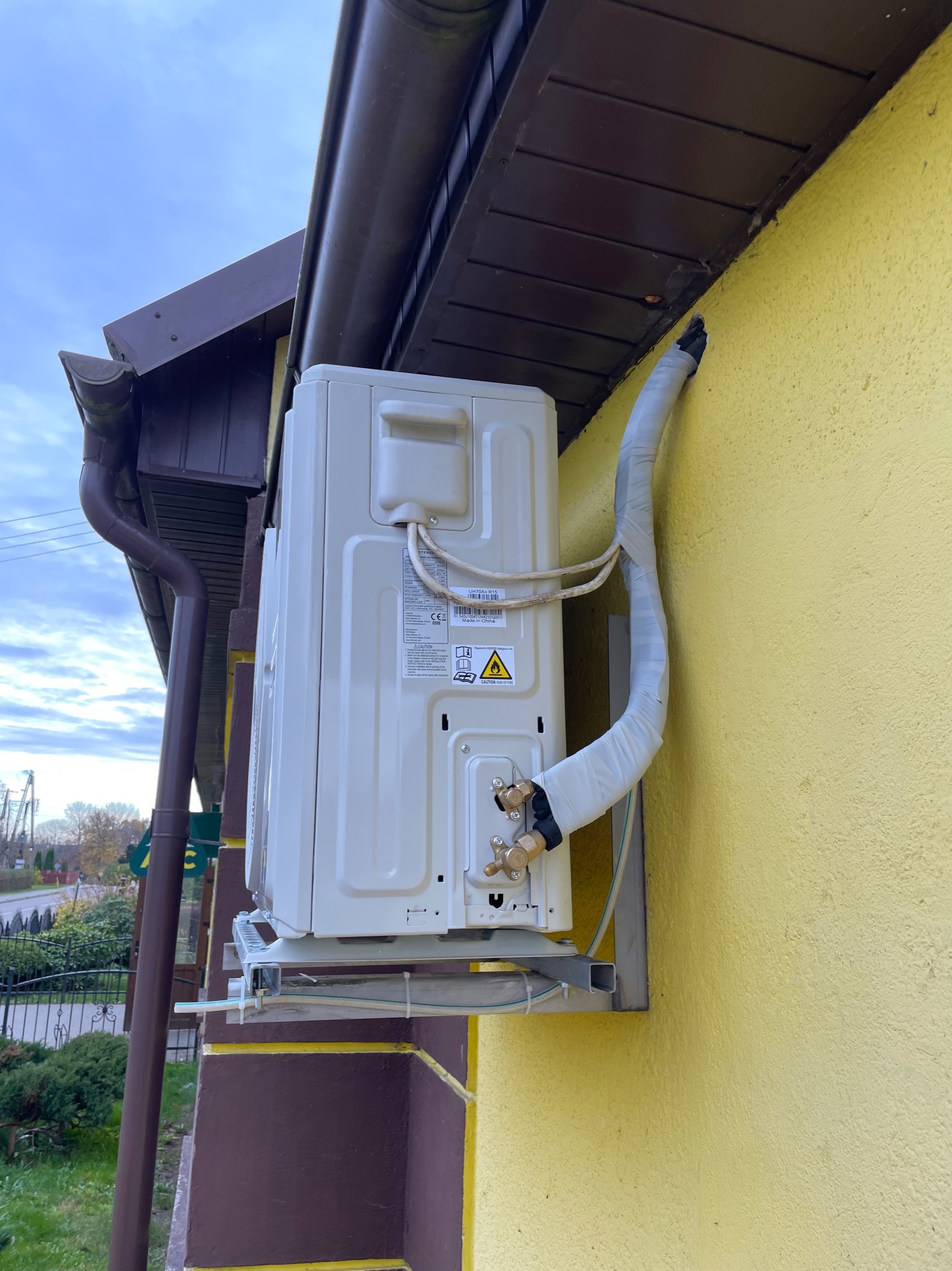 Zamontowana jednostka zewnętrzna klimatyzacji na żółtej ścianie budynku, widoczne rury w izolacji i elementy mocujące. Ujęcie z dołu, fragment dachu i rynny.