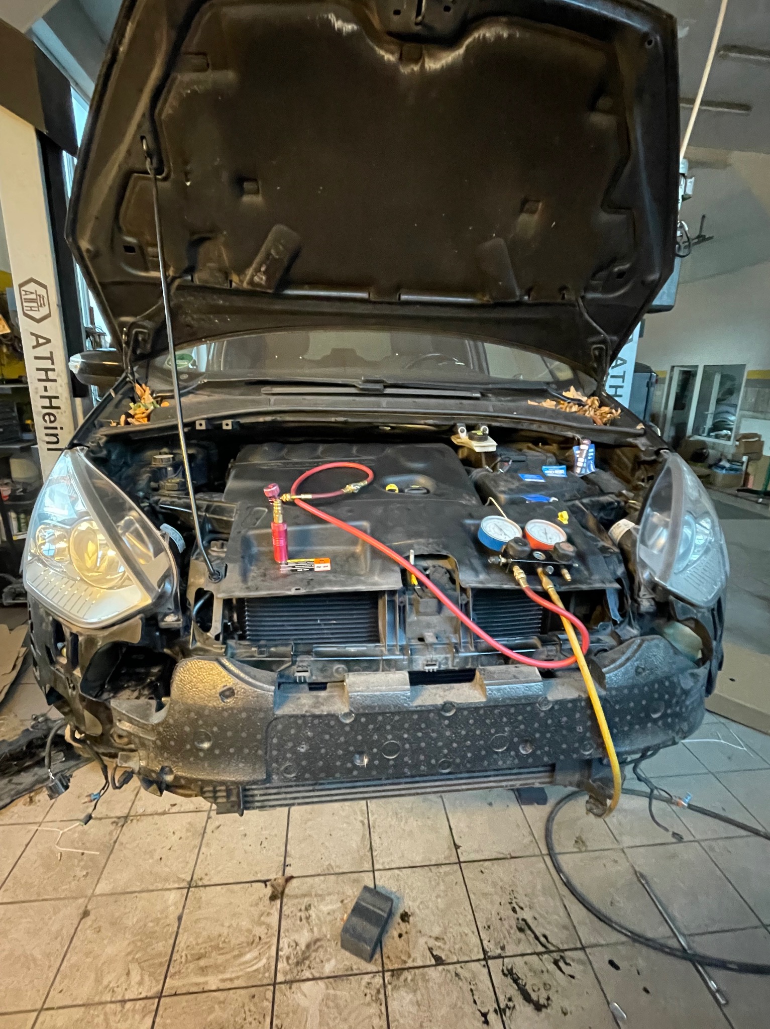 Otwarta maska samochodu z podłączonym sprzętem diagnostycznym klimatyzacji. Widoczne przewody i manometry. Warsztatowy klimat, płytki na podłodze.