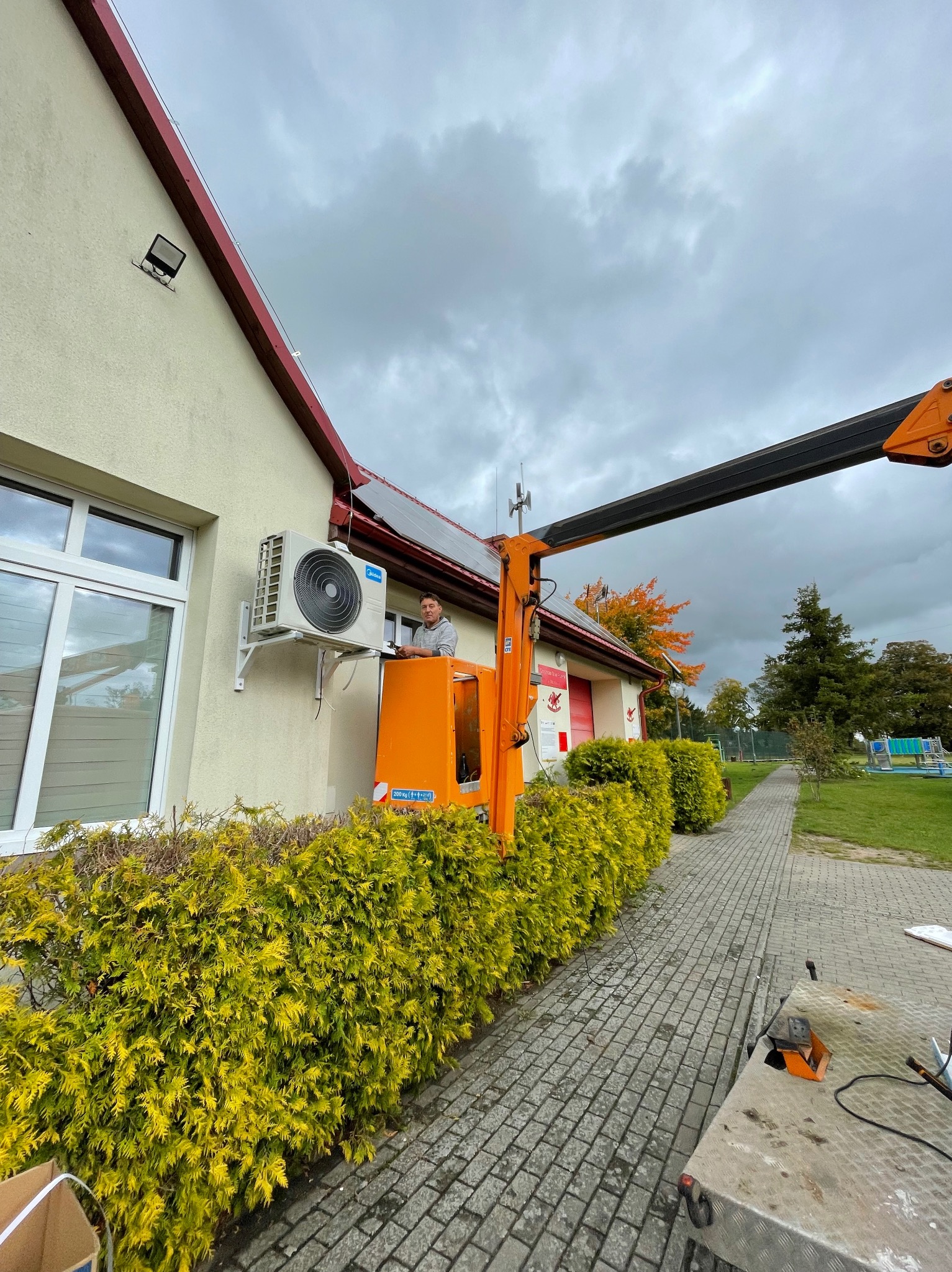 Montaż klimatyzacji zewnętrznej na budynku w Bytowie. Pracownik na podnośniku instaluje jednostkę. Widoczne krzewy i chodnik.