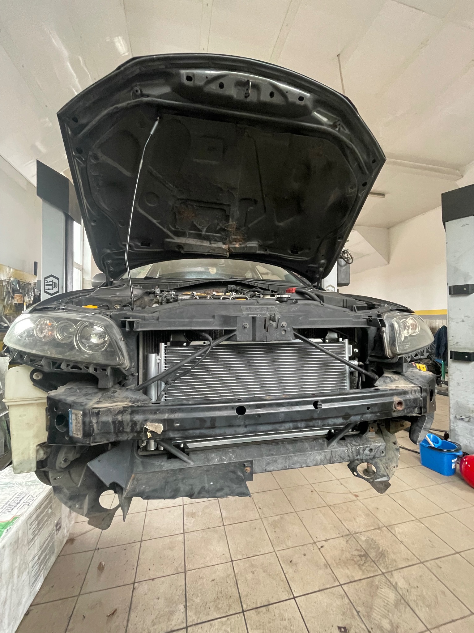 Samochód z otwartą maską w warsztacie, widoczny wymieniony radiator klimatyzacji. Detal: niebieski pojemnik i czerwona butla w tle po prawej stronie.