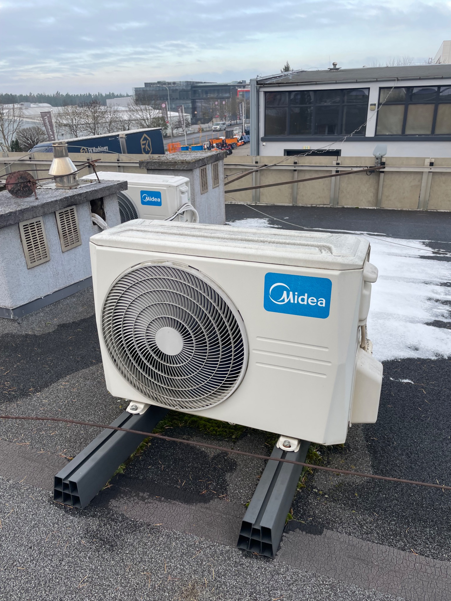 Biała jednostka zewnętrzna klimatyzacji Midea na dachu budynku, zamontowana na szarych wspornikach. W tle zabudowa miejska i pochmurne niebo.