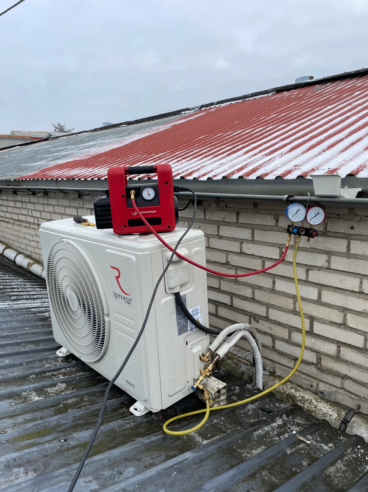 Biała jednostka zewnętrzna klimatyzacji Rotenso na dachu, podłączona do czerwonej pompy próżniowej i manometrów. Widoczny ceglany komin i ośnieżony dach.