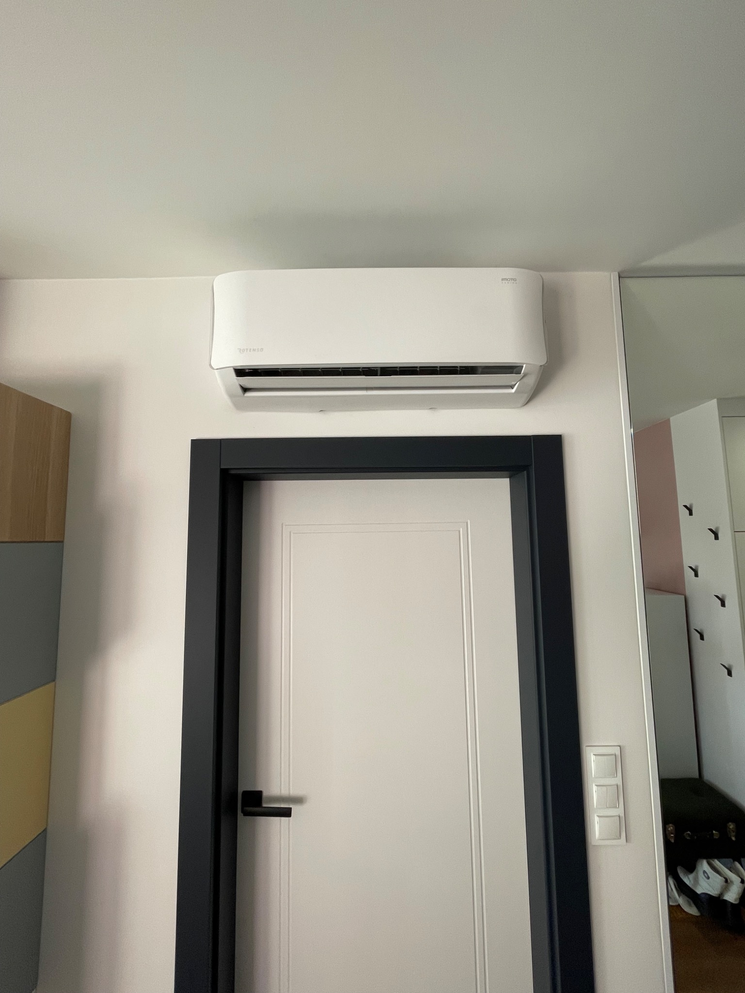 Biały klimatyzator Rotenso zamontowany nad drzwiami z czarną ościeżnicą w jasnym wnętrzu. Widoczne elementy szafy i włącznik światła. Minimalistyczny design.