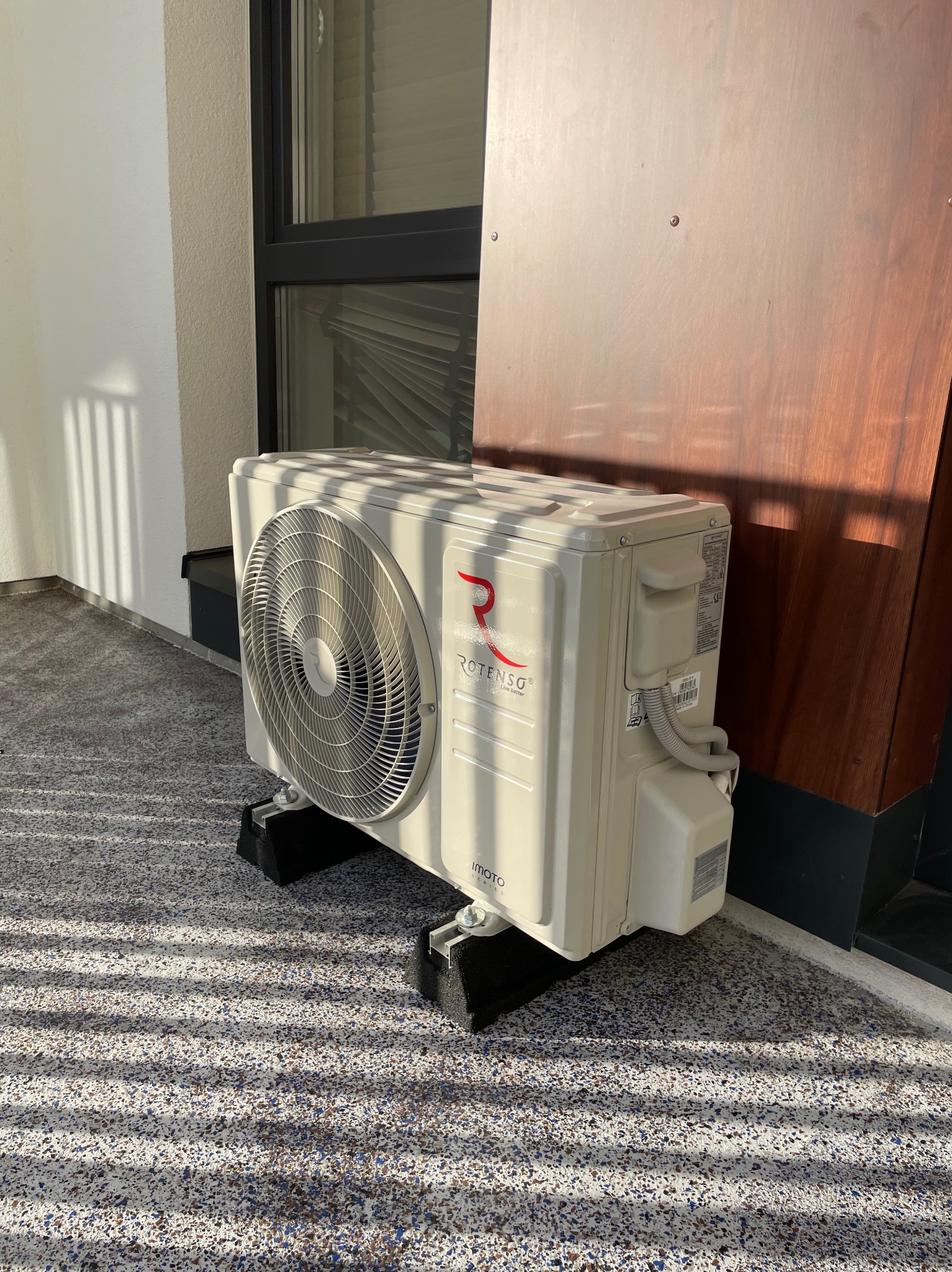 Biała jednostka zewnętrzna klimatyzacji Rotenso Imoto na balkonie, z widocznymi cieniami i elementami elewacji. Ujęcie z bliska, prezentujące detale urządzenia.