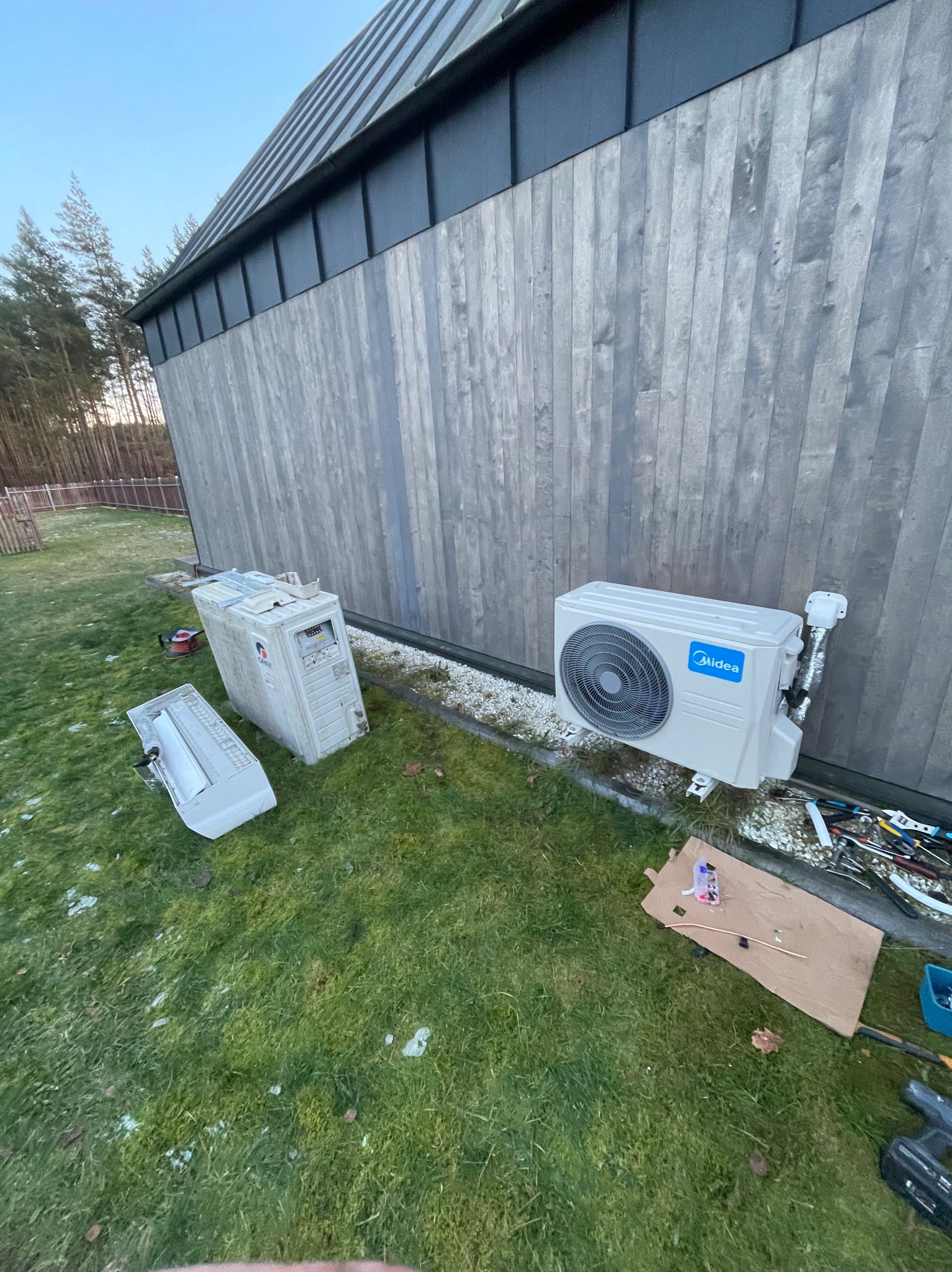Montaż klimatyzacji Midea na elewacji domu. Obok leżą zdemontowane elementy starego urządzenia i narzędzia na trawie. Widok z boku, lekki skos.