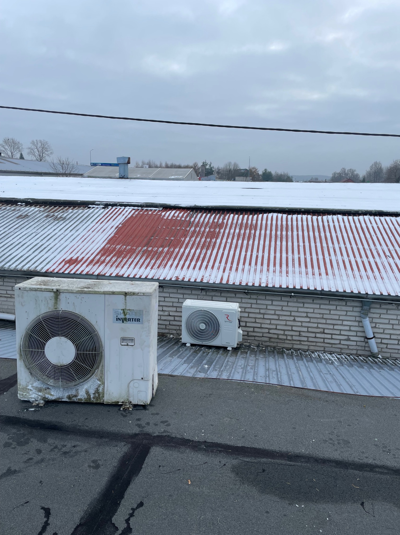 Dwa klimatyzatory na dachu budynku w zimowej scenerii. Jeden z nich jest wyraźnie zabrudzony, drugi nowszy. W tle widać budynki pokryte śniegiem i pochmurne niebo.