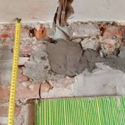 Nierówna ściana z cegły po kuciu, częściowo pokryta zaprawą. Widoczne kable elektryczne i miara zwinięta. Prace remontowe w trakcie, surowy stan.