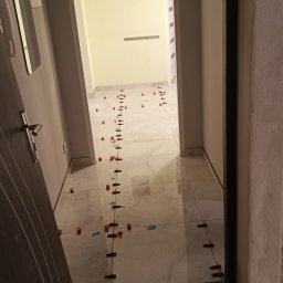 RemontLine - Układanie jasnych płytek podłogowych w korytarzu z użyciem systemu poziomowania, widoczne krzyżyki dystansowe i kliny. W tle obrazek z motywem kawy.