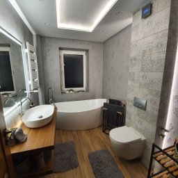 RemontLine - Nowoczesna łazienka z owalną wanną, drewnianym blatem pod umywalką i szarą mozaiką na ścianie. Oświetlenie LED podkreśla elegancki wystrój.