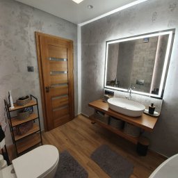 RemontLine - Stylowa łazienka z drewnianymi akcentami: dębowe drzwi, umywalka na blacie z półkami, lustro z podświetleniem LED i dekoracyjna ściana w odcieniach szarości.