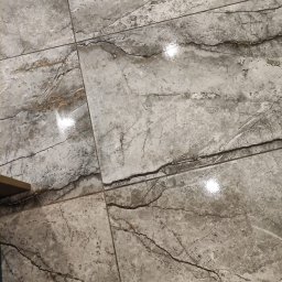 RemontLine - Szare płytki podłogowe imitujące marmur, ułożone równo z widocznymi fugami. Powierzchnia błyszcząca, odbijająca światło. Widok z góry, fragment podłogi.