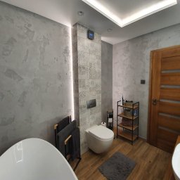 RemontLine - Nowoczesna łazienka z szarą, teksturowaną ścianą, drewnianymi drzwiami i elementami w stylu boho. Uwagę zwraca podświetlany pionowy panel z dekoracyjnymi płytkami.