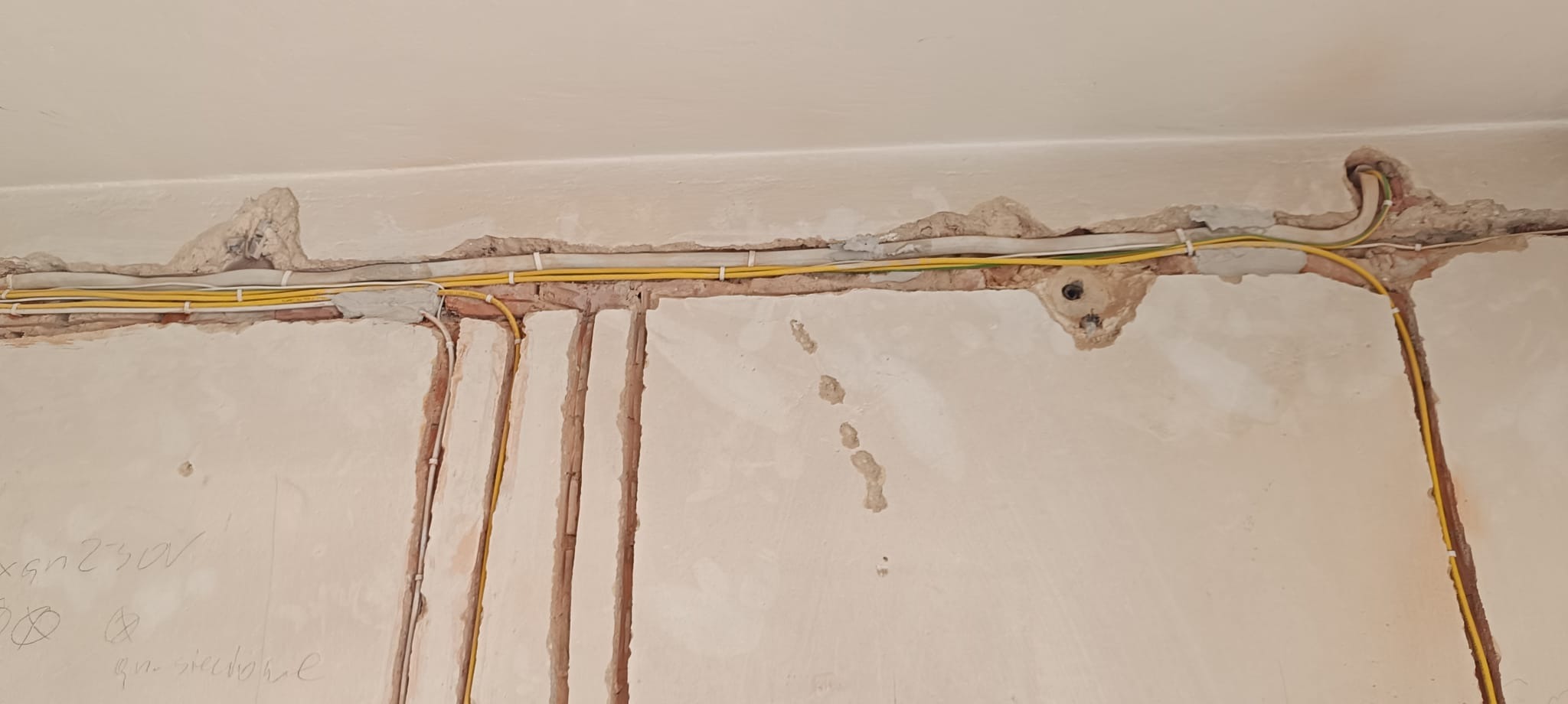 Żółte kable elektryczne ułożone w bruzdach na ścianie, zabezpieczone białymi uchwytami. Ściana w trakcie remontu, widoczne ślady po kuciu.