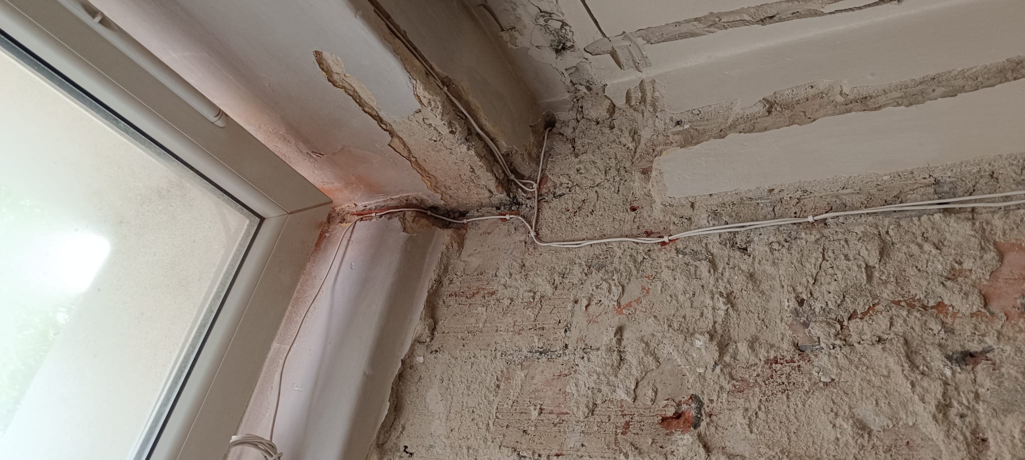 Fragment ściany z widocznymi przewodami elektrycznymi, surowym tynkiem i narożnikiem okna. Widoczne ślady po kuciu i przygotowaniu do dalszych prac wykończeniowych.