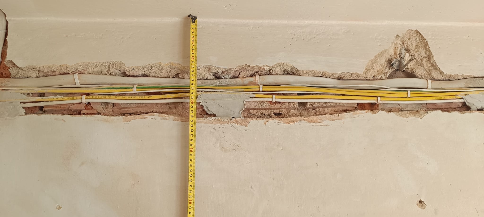 Żółte i białe przewody elektryczne ułożone w bruzdach w ścianie, częściowo zakryte zaprawą murarską, z widoczną taśmą mierniczą.