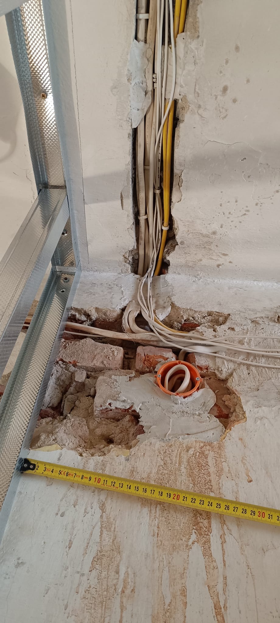 Przygotowanie do montażu instalacji elektrycznej w ścianie z cegły, widoczne przewody, puszka instalacyjna i metalowy profil sufitowy. Prace remontowe w toku.