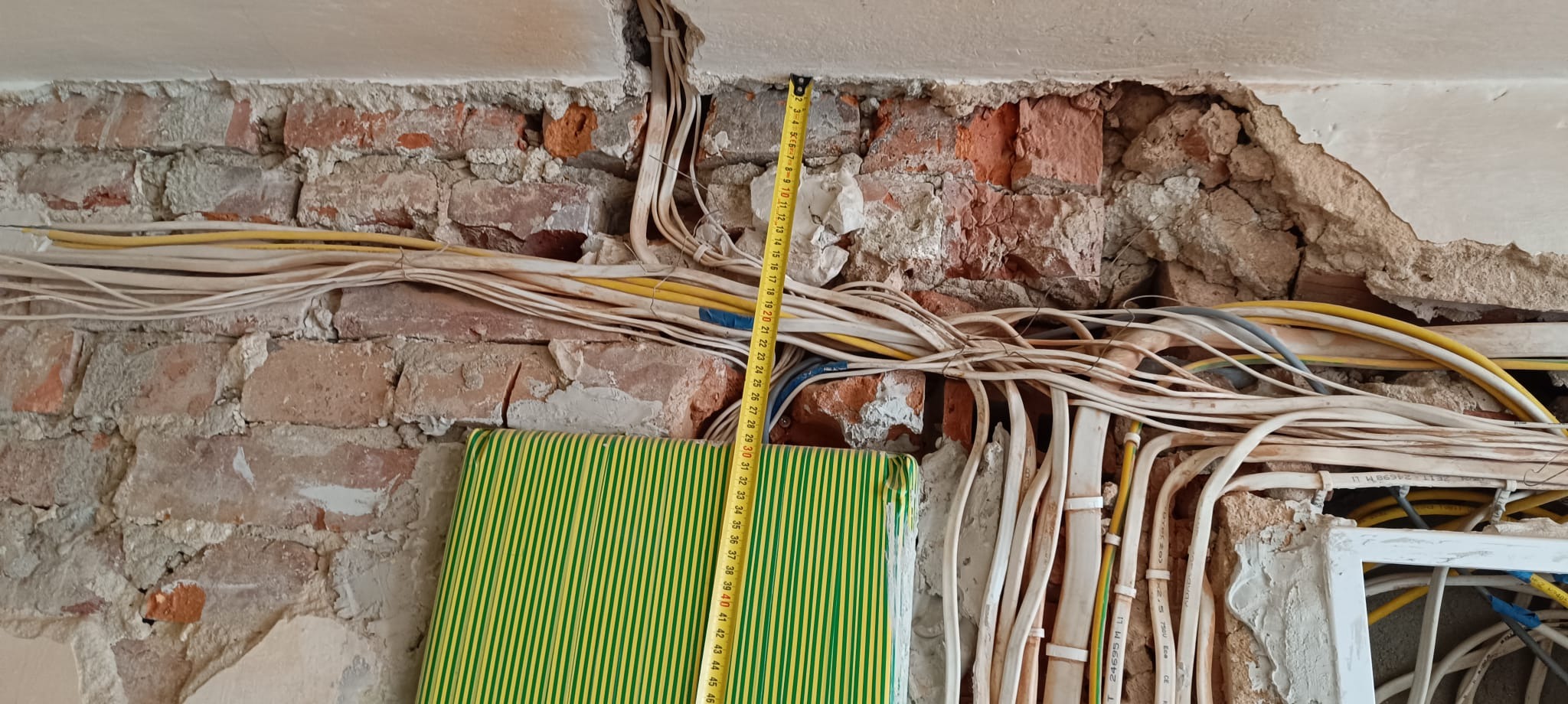 Ściana z odsłoniętą instalacją elektryczną w trakcie remontu, widoczne przewody, cegły i częściowo zielony element, pomiar odległości żółtą miarką.
